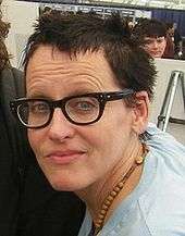 A photograph of a middle-aged woman with short, messy hair, wearing glasses and a light blue T-shirt