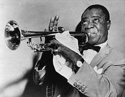 Short-haired black man in his fifties blowing into a trumpet. He is wearing a light-colored sport coat, a white shirt and a bow tie. He is faced left with his eyes looking upwards. His right hand is fingering the trumpet, with the index finger down and three fingers pointing upwards. The man's left hand is mostly covered with a handkerchief and it has a shining ring on the little finger. He is wearing a wristwatch on the left wrist.