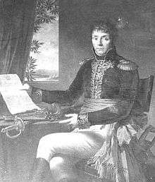 Black and white print of a man in dark military coat with gold braid and white breeches. Seated at a table, he gestures with his left hand toward a document held in his right hand.