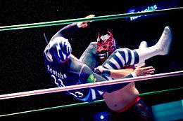 A color photograph of a wrestler in black and white gear, including a black and white mask executing a head scissors takedown on a wrestler in black and red mask with ornamental horns.