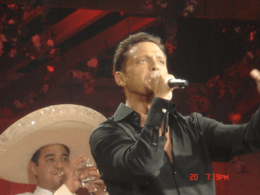 A man in the front singing to a microphone and on the back another man wearing a charro suit and playing the trumpet.