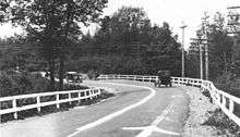 An old photograph of a bend in a road surrounded by trees and power poles. There are wooded guardrails on either side of the road, with a white-painted centerline separating the two lanes of traffic. Two old cars are approaching the curve which also has arrows to denote the direction of traffic.