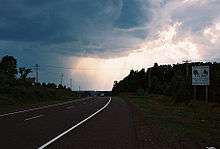 a highway curving over the crest of a hill flanked by telephone poles on either side. There is a white road sign on the right side of the road and storm clouds in the sky.