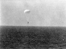 A conical black spacecraft falling towards the surface of the ocean under a single white parachute, seen from some distance away. Very little detail can be seen.