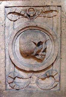 Close-up of a rectangular-shaped carving in stone. In the centre of the rectangle is a circle representing a mirror, and within the circle is a grinning skull. The circle is framed by ram's horns.