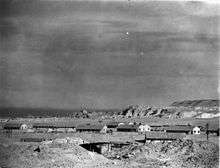 A black and white photograph of a desert landscape, with huts in the mid-distance and the walls of a city in the far distance