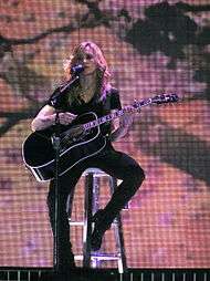 Faraway image of a blond woman singing in front of a bloomy backdrop. She is wearing a black blouse and pants of the same color. She is holding a black guitar and a microphone to her mouth.
