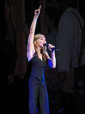 A blond woman singing into a microphone in her left hand, while pointing her right hand upwards. She is dressed in black trousers with a black, sleeveless jacket. Her hair falls in curls around her, but is set in place by a black band.