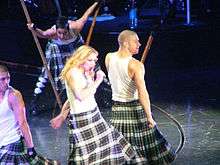 A blond woman with long wavy hair singing on stage, wearing a white T-shirt and a black-and-white patterned skirt, holding a microphone in her hand. She is surrounded by dancers in similar attire, carrying long poles.