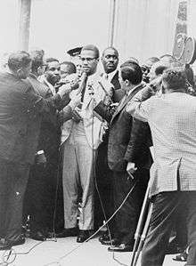 Malcolm X is surrounded by reporters with microphones, while a television camera captures the scene