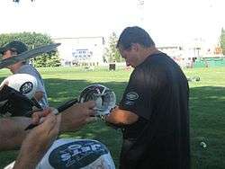 Waist up photograph from the side of Mangini wearing a green New York Jets t-shirt and signing an autograph