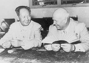 A balding Russian man (Nikita Kruschev) and a younger Chinese man (Mao Zedong) sit and smile, the balding man holding a fan