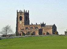 A sandstone church seen from the south at a distance standing on a mound. The tower to the left has crocketted pinnacles and the nave and aisle are crenellated. The foreground consists of a grassy field