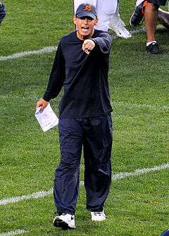 Color photograph of Trestman wearing a dark blue long-sleeve t-shirt, dark blue warmup pants and a blue hat with a Chicago Bears logo pointing with his left hand and holding a sheet of paper in his right hand at his side