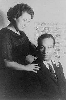 Black and white image of a woman in a dress standing behind a man sitting in a chair. He is wearing a suit and her arms are placed on his shoulders.