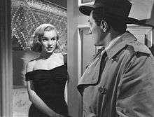 Monroe in The Asphalt Jungle. She is wearing a black dress and stands in a doorway, facing a man wearing a trench coat and a fedora