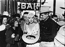 Monroe in The Misfits, holding a wide-brimmed hat filled with dollar bills and standing next to Clark Gable and Thelma Ritter. Behind them is a sign spelling "BAR" and a crowd of people.