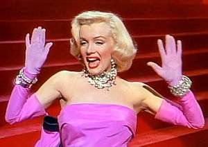 Bust of a blond woman in short curled hair and wearing a bright pink, sleeveless dress. Putting both her hands up, she looks to the right of the image.