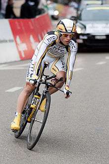A road racing cyclist wearing a yellow and white jersey with black trim, a matching helmet, and sunglasses. A car is partly visible behind him, as are roadside barricades.