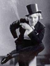 Black-and-white image of a young woman wearing a tuxedo-like suit and top hat. She sits with her right leg, almost parallel to the ground, over her left knee. She leans forward and gazes out directly with a tight-lipped smile. In her right hand she holds a cigarette from which a large plume of smoke rises.