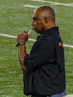 Color photograph of African-American man (Marvin Lewis) wearing black sport shirt, standing on football sideline and holding a capped Sharpie marker to his lips.