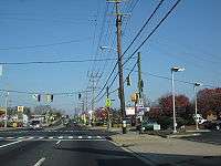 A four lane divided highway in a commercial area at a traffic light. On the traffic light pole, a Maryland Route 171 shield with a right arrow can be seen.