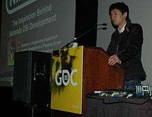 A man stands behind a podium, talking to the audience. Next to him is a projection screen displaying a presentation program slide.