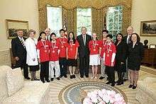 Fourteen people stand in a line, seven of whom are wearing medals and red T-shirts, and the other seven of whom are dressed formally.