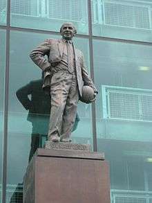 A bronze statue of a bald man wearing a suit, with his hand on his right hip and holding a football to his left hip.