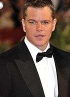 Matt Damon, a caucasian male in his late-30s with dark hair, looks into the camera. He wears a black suit and white shirt with a black bow-tie.