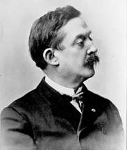 Profile of a white man with a mustache, wearing a dark suit coat and bow tie.