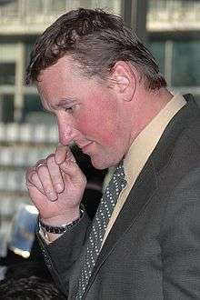 Matthew Pinsent is pictured side on. He is wearing a brownish suit and has one hand up to his face
