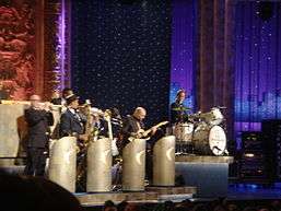 Several men are playing instruments, including trumpet, saxophone, trombone, and guitar, behind bandstands; at a higher elevation to the right of them is another man behind a drum kit and looking over at the band members; the bass drum is labeled "The Max Weinberg 7"; behind all of them is a paster sculpture wall and a series of vertical curtains, some translucent with a painted cityscape behind them