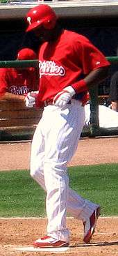 A dark-skinned man in a red baseball jersey, white baseball pants with red pinstripes, and a red batting helmet steps on home plate with his left foot while looking down.