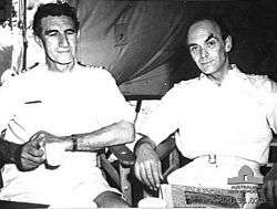 An informal photograph featuring two men sitting beside each other on wooden chairs. Both are wearing white naval uniforms and appear to be in a tent. The man on the left has dark, slightly curly hair, his arms are semi-crossed with a mug in his right hand and a watch on his left wrist. He is not looking at the camera. The other man has short hair and is slightly balding. He has his arms resting on the chair's arm rests and is looking at the camera.