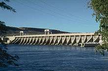 A concrete hydroelectric dam's spillway on the downriver side. Water is in the foreground and power lines run overhead.