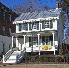 A two-story white wooden house with decorative touches, a pointed metal roof and green shutters. A sign in the center of the porch reads "Irvington History Center" while another at the right on the story below says "Frocks, Frills and Furbelous 1760-2009"