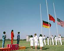 On a grassed outdoor field, three women stand on a podium to the left of the shot, while people wearing all white raise three flags on flagpoles situated at the right of shot.