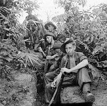 Six soldiers sit in a clearing in the jungle, several of whom are smoking. The man in the foreground stares towards the camera.