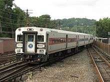 A train headed by a light gray car with a red stripe on the side on a track between two others in a walled area moving from right to left across the image. On its front is the number 6222.