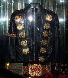 A black jacket with five round golden medals on its left and right shoulders, a gold band on its left arm sleeve, and two belt straps on the right bottom sleeve. Underneath the jacket is a golden belt, with a round ornament in its center.