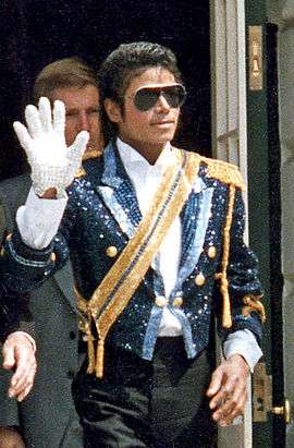A mid-twenties Michael Jackson wearing a sequined military jacket and dark sunglasses. He is walking while waving his right hand, which is adorned with a white glove. His left hand is bare.
