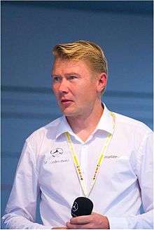 Head and shoulders of a man in his forties with blonde hair and grey eyes. He is wearing a white shirt which bears the Mercedes-Benz and AMG logos, and is holding a microphone in both of his hands.