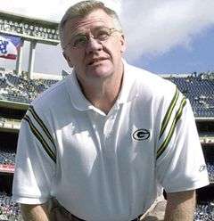 Candid waist-up photograph of Mike Sherman in a football stadium wearing a white Green Bay Packers polo shirt