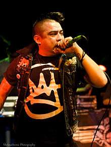 A man with a mohawk in a black T-shirt and open black jacket holds a microphone close to his mouth
