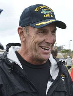 Color head-and-shoulders photograph of a smiling white man (Mike Mularkey) wearing a black Jacksonville Jaguars jacket and a navy blue USS Bataan baseball cap.