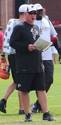Candid photograph of Smith standing on a football field with a whistle in his mouth holding a pen and a laminated sheet of paper in his right hand and wearing black shorts, a black long-sleeved t-shirt and a white baseball cap all of which bear the logo of the Atlanta Falcons