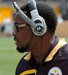 Color head shot of African-American man (Mike Tomlin) in profile on a football sideline wearing a black and gold Pittsburgh Steelers jacket, Motorola headset and sunglasses.
