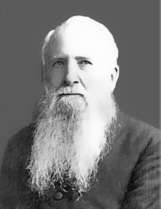 Head and shoulders of an older white man with a long beard and white hair, wearing a dark suit coat.