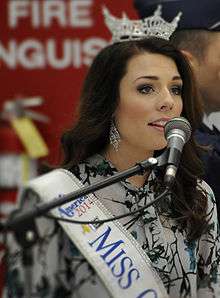 A young woman with brown hair, wearing a crown on her head and a Miss Oregon sash over her dress, singing into a microphone